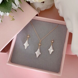 May include: A set of pearl and gold earrings and a necklace in a pink box. The earrings and necklace feature a white pearl in the shape of a star with a small gold diamond in the center.