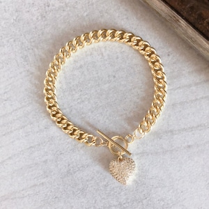 May include: A gold chain bracelet with a toggle clasp and a heart-shaped charm encrusted with clear rhinestones.