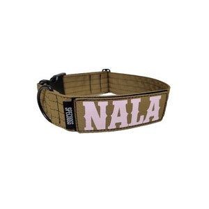 Pode incluir: Uma coleira de cachorro marrom com uma fivela preta e um remendo rosa que diz "NALA" em letras brancas. A coleira é feita pela SPECDOGS.