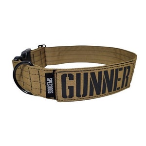 May include: A tan nylon dog collar with black stitching and a black buckle. The collar has a black patch with the word "GUNNER" printed in white letters and the word "SPECDOGS" printed in black letters.