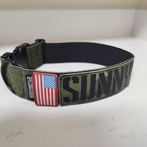 Pode incluir: Coleira camuflada verde para cães com o nome "SUNNY" em letras pretas. A coleira apresenta uma fivela preta e um patch com a bandeira americana. A marca "SPECDOGS" também é visível.