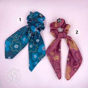 May include: Two colorful hair scrunchies with matching fabric ties. The scrunchies are made from recycled material and feature a floral pattern. The scrunchie on the left is blue and green, while the scrunchie on the right is red and orange.
