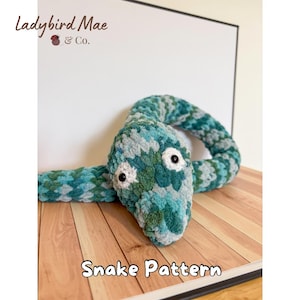 May include: A plush, crocheted snake in shades of teal, green, and gray. The snake has large white eyes with black pupils and is positioned on a wooden surface. The text "Snake Pattern" is visible below the snake.