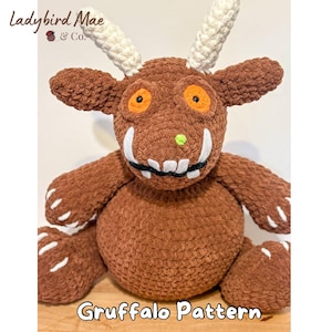 Peut inclure: Peluche Gruffalo marron crochetée avec des cornes blanches, des yeux orange et un nez vert. La peluche a des griffes et des dents blanches. Le texte "Gruffalo Pattern" est en bas de l'image.