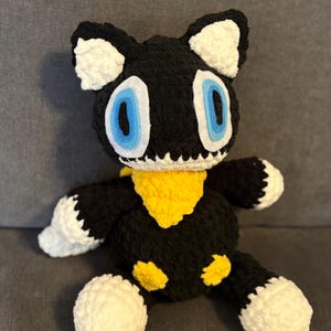 May include: A handmade crochet plush toy of a black cat with white accents on the ears, paws, and tail. The cat has large blue eyes and a yellow scarf. The plush is sitting on a gray surface.