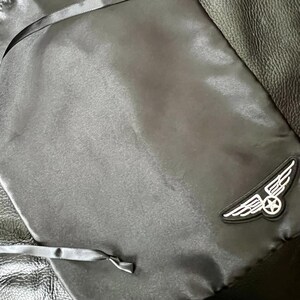 Puede incluir: Bolsa negra con cordón y acabado satinado. La bolsa presenta un panel de cuero y un parche con un emblema alado blanco y una estrella. El cordón es una cinta fina y negra.