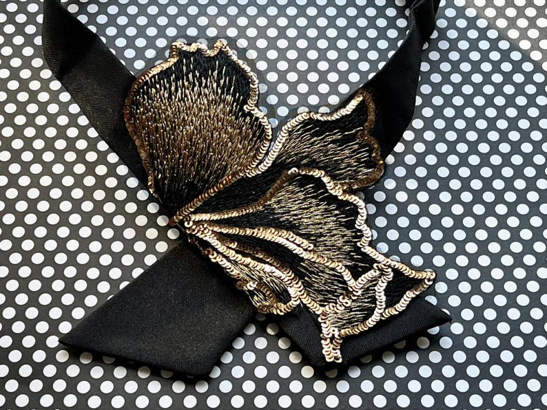 Puede incluir: Una pajarita negra con un gran dise&ntilde;o de mariposa bordada. La mariposa presenta lentejuelas y hilo dorado, creando un efecto brillante. La pajarita est&aacute; sobre un fondo de lunares blancos y negros.