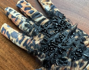 Puede incluir: Primer plano de un guante con estampado de leopardo y adornos de cuentas negras. El guante presenta un ribete de encaje negro transparente y está hecho de una tela suave estampada. Los dedos son visibles, mostrando el intrincado diseño.