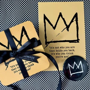 May include: A gift set featuring a black and white crown design. The set includes a gift tag tied with a black ribbon, a greeting card, and a button. The tag and card have the quote, "It's not who you are that holds you back, it's who you think you're not."