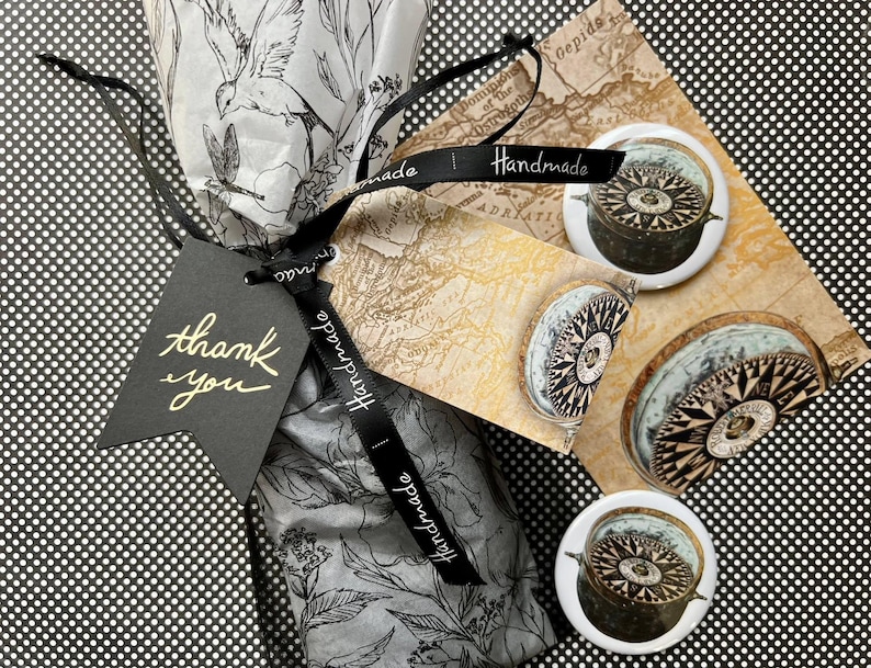 K&ouml;nnte beinhalten: Draufsicht auf ein Geschenk, das in florales Seidenpapier verpackt und mit einem schwarzen Band mit der Aufschrift "Handmade" gebunden ist. Ein schwarzes Etikett tr&auml;gt die Aufschrift "thank you". Drei Pins mit Kompassmotiven liegen auf einer kartenbedruckten Oberfl&auml;che und zeigen nautisches Design.