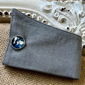 Puede incluir: Un puño de camisa gris a cuadros con un gemelo redondo plateado. El gemelo presenta un diseño de mapa del mundo azul y blanco. El puño está doblado, mostrando el gemelo. El fondo incluye un marco blanco decorativo.
