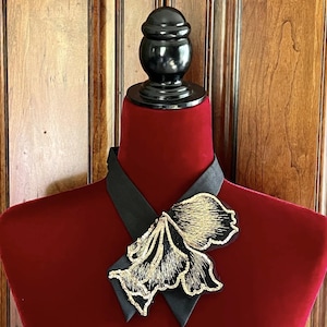 Puede incluir: Broche floral negro y dorado sobre un cuello negro, exhibido en un maniqu&iacute; de terciopelo rojo. El broche presenta un intrincado bordado dorado y un contorno negro. El fondo es una puerta de madera.