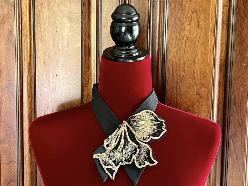 Puede incluir: Broche floral negro y dorado sobre un cuello negro, exhibido en un maniqu&iacute; de terciopelo rojo. El broche presenta un intrincado bordado dorado y un contorno negro. El fondo es una puerta de madera.