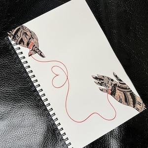 May include: A white spiral-bound notebook with a drawing of two hands connected by a red string in a heart shape. The hands are decorated with detailed black and brown patterns. The notebook is on a black textured surface.