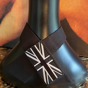 K&ouml;nnte beinhalten: Ein schwarzes Nackenband mit einem Union Jack-Aufn&auml;her. Der Aufn&auml;her zeigt ein wei&szlig;es und schwarzes Design der britischen Flagge. Das Nackenband ist um einen schwarzen Schaufensterkopf gewickelt.