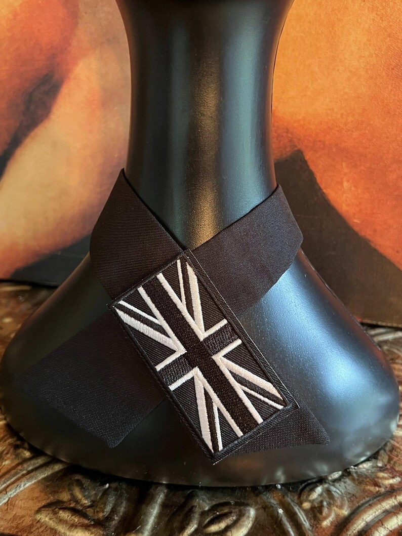 K&ouml;nnte beinhalten: Ein schwarzes Nackenband mit einem Union Jack-Aufn&auml;her. Der Aufn&auml;her zeigt ein wei&szlig;es und schwarzes Design der britischen Flagge. Das Nackenband ist um einen schwarzen Schaufensterkopf gewickelt.