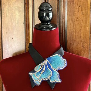 Puede incluir: Un collar decorativo exhibido en un maniqu&iacute; rojo. El collar presenta una banda de sat&eacute;n negro y una gran flor azul bordada con detalles dorados. El maniqu&iacute; se encuentra sobre un fondo de paneles de madera.