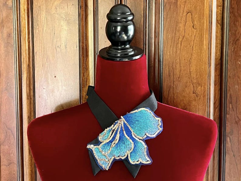 Puede incluir: Un collar decorativo exhibido en un maniqu&iacute; rojo. El collar presenta una banda de sat&eacute;n negro y una gran flor azul bordada con detalles dorados. El maniqu&iacute; se encuentra sobre un fondo de paneles de madera.
