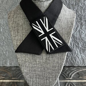 K&ouml;nnte beinhalten: Eine schwarze Krawatte mit einem Union-Jack-Aufn&auml;her. Die Krawatte wird auf einem grauen Mannequinkopf pr&auml;sentiert. Der rechteckige Aufn&auml;her hat einen schwarzen Hintergrund und ein wei&szlig;es Design. Die Krawatte ist aus einem glatten, dunklen Stoff gefertigt.