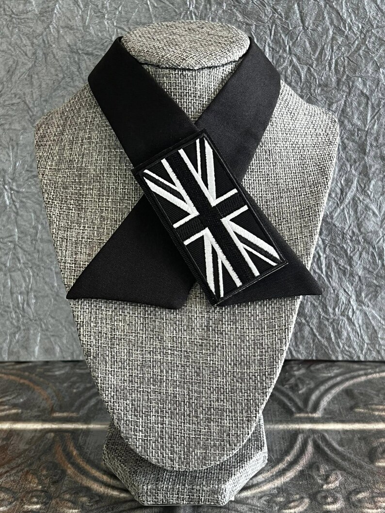 K&ouml;nnte beinhalten: Eine schwarze Krawatte mit einem Union-Jack-Aufn&auml;her. Die Krawatte wird auf einem grauen Mannequinkopf pr&auml;sentiert. Der rechteckige Aufn&auml;her hat einen schwarzen Hintergrund und ein wei&szlig;es Design. Die Krawatte ist aus einem glatten, dunklen Stoff gefertigt.