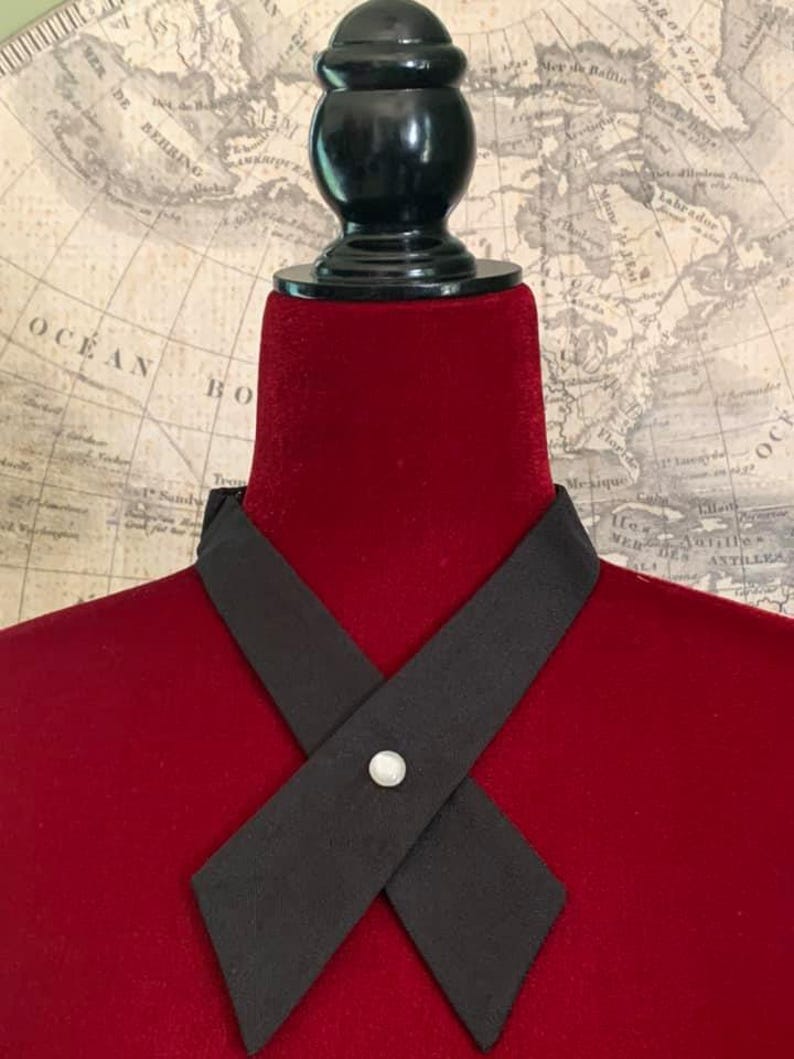 Puede incluir: Una corbata negra con un dise&ntilde;o en forma de X, asegurada con un peque&ntilde;o bot&oacute;n redondo con forma de perla. La corbata se exhibe en un maniqu&iacute; de terciopelo rojo, con un mapa antiguo de fondo. El maniqu&iacute; tiene una parte superior negra y redondeada.