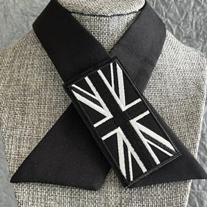 K&ouml;nnte beinhalten: Eine schwarze Krawatte mit einem rechteckigen Aufn&auml;her mit einem wei&szlig;-schwarzen Union-Jack-Flaggen-Design. Die Krawatte ist &uuml;ber einem Mannequinkopf mit einem strukturierten, grauen Stoff drapiert.