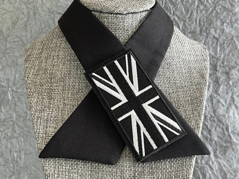 K&ouml;nnte beinhalten: Eine schwarze Krawatte mit einem rechteckigen Aufn&auml;her mit einem wei&szlig;-schwarzen Union-Jack-Flaggen-Design. Die Krawatte ist &uuml;ber einem Mannequinkopf mit einem strukturierten, grauen Stoff drapiert.