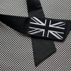K&ouml;nnte beinhalten: Eine schwarze Krawatte mit einem rechteckigen Aufn&auml;her mit der Union-Jack-Flagge in Wei&szlig; und Grau. Die Krawatte ist aus glattem, mattem Stoff gefertigt und vor einem schwarz-wei&szlig;en Tupfenhintergrund platziert.