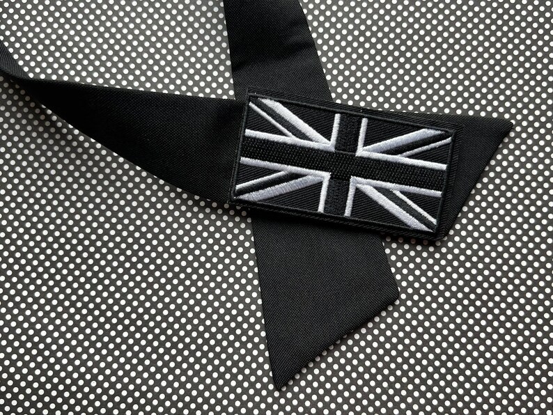 K&ouml;nnte beinhalten: Eine schwarze Krawatte mit einem rechteckigen Aufn&auml;her mit der Union-Jack-Flagge in Wei&szlig; und Grau. Die Krawatte ist aus glattem, mattem Stoff gefertigt und vor einem schwarz-wei&szlig;en Tupfenhintergrund platziert.