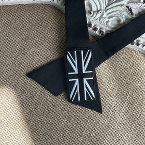 K&ouml;nnte beinhalten: Schwarze Krawatte mit Union-Jack-Aufn&auml;her. Die Krawatte besteht aus einem glatten, dunklen Stoff und hat einen rechteckigen Aufn&auml;her mit dem wei&szlig;-blauen Design der britischen Flagge. Die Krawatte wird auf einer strukturierten, neutral get&ouml;nten Oberfl&auml;che pr&auml;sentiert.