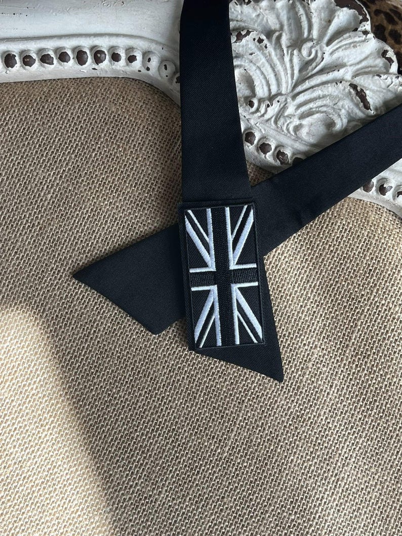 K&ouml;nnte beinhalten: Schwarze Krawatte mit Union-Jack-Aufn&auml;her. Die Krawatte besteht aus einem glatten, dunklen Stoff und hat einen rechteckigen Aufn&auml;her mit dem wei&szlig;-blauen Design der britischen Flagge. Die Krawatte wird auf einer strukturierten, neutral get&ouml;nten Oberfl&auml;che pr&auml;sentiert.