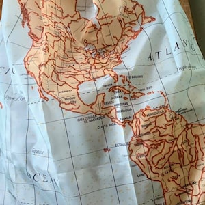 Könnte beinhalten: Eine Vintage-Weltkarte, gedruckt auf einem hellblauen Stoff mit orangefarbenen und braunen Rändern. Die Karte zeigt die Kontinente Nord- und Südamerika mit dem Atlantik rechts.