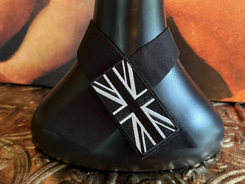 K&ouml;nnte beinhalten: Schwarzes Dekorationsobjekt mit einem schwarz-wei&szlig;en Union-Jack-Patch. Der rechteckige Patch ist auf einem schwarzen Stoffband angebracht, das um ein schwarzes, abgerundetes Objekt gewickelt ist. Der Hintergrund ist ein verschwommener Farbmix.