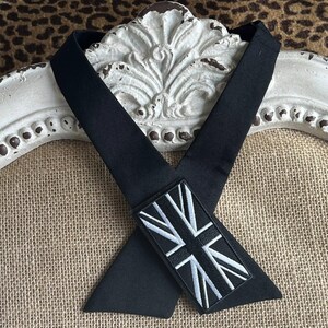 K&ouml;nnte beinhalten: Schwarzes Nackenband mit einem rechteckigen Aufn&auml;her mit einem wei&szlig;-grauen Union Jack-Flaggen-Design. Das Nackenband ist aus einem glatten, dunklen Stoff gefertigt und wird vor einem dekorativen wei&szlig;en Hintergrund pr&auml;sentiert.