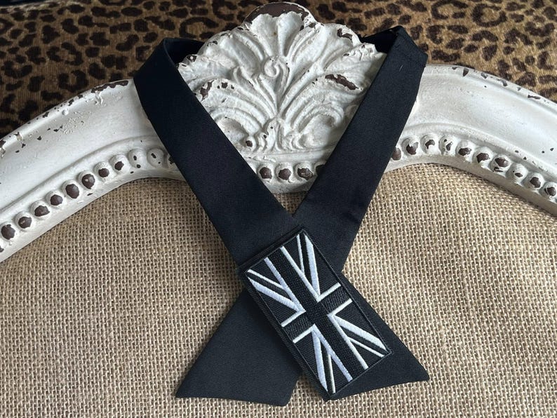 K&ouml;nnte beinhalten: Schwarzes Nackenband mit einem rechteckigen Aufn&auml;her mit einem wei&szlig;-grauen Union Jack-Flaggen-Design. Das Nackenband ist aus einem glatten, dunklen Stoff gefertigt und wird vor einem dekorativen wei&szlig;en Hintergrund pr&auml;sentiert.