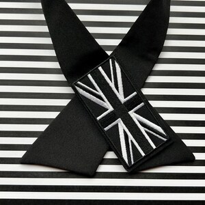 K&ouml;nnte beinhalten: Schwarze Hosentr&auml;ger mit einem Union-Jack-Aufn&auml;her. Die Hosentr&auml;ger sind gekreuzt und liegen auf einem schwarz-wei&szlig; gestreiften Hintergrund. Der Aufn&auml;her zeigt eine wei&szlig;-schwarz gestickte britische Flagge.