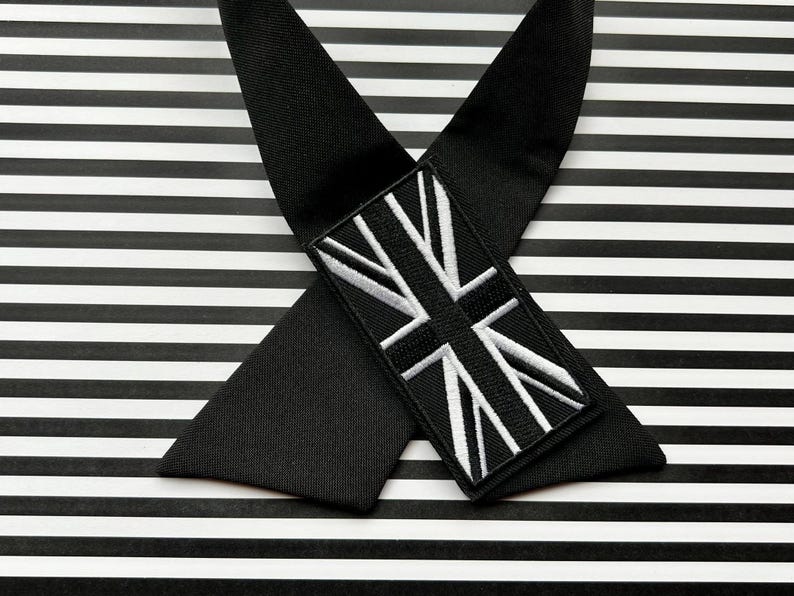 K&ouml;nnte beinhalten: Schwarze Hosentr&auml;ger mit einem Union-Jack-Aufn&auml;her. Die Hosentr&auml;ger sind gekreuzt und liegen auf einem schwarz-wei&szlig; gestreiften Hintergrund. Der Aufn&auml;her zeigt eine wei&szlig;-schwarz gestickte britische Flagge.