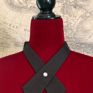 K&ouml;nnte beinhalten: Eine schwarze Krawatte in X-Form, befestigt mit einem perlenartigen Knopf. Die Krawatte wird auf einem roten Samt-Mannequinkopf mit schwarzem St&auml;nder pr&auml;sentiert. Im Hintergrund ist eine Vintage-Karte zu sehen.