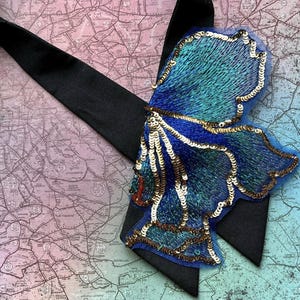 Puede incluir: Una pajarita negra con un adorno grande en forma de mariposa azul y turquesa. La mariposa est&aacute; adornada con lentejuelas doradas y bordados intrincados. El fondo es un mapa.