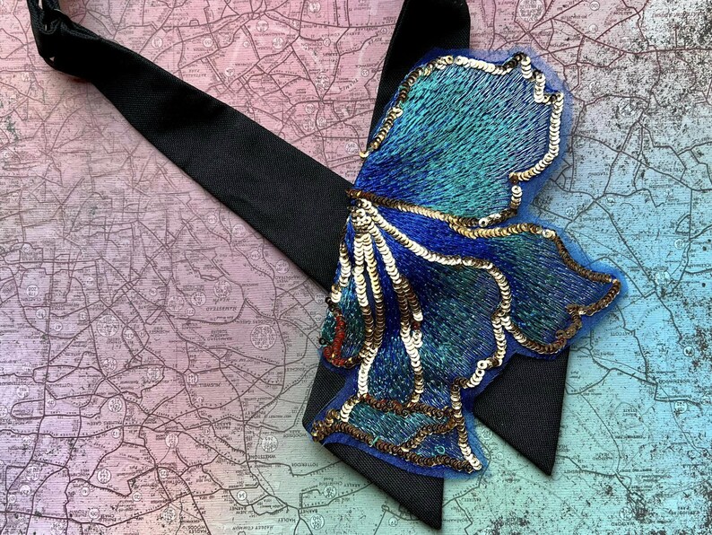 Puede incluir: Una pajarita negra con un adorno grande en forma de mariposa azul y turquesa. La mariposa est&aacute; adornada con lentejuelas doradas y bordados intrincados. El fondo es un mapa.
