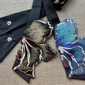 Puede incluir: Dos adornos decorativos con forma de mariposa con lazos de cinta negra. Una mariposa est&aacute; bordada con lentejuelas doradas, marrones y rojas. La otra es azul y turquesa con lentejuelas plateadas. Las cintas tienen cierres a presi&oacute;n.