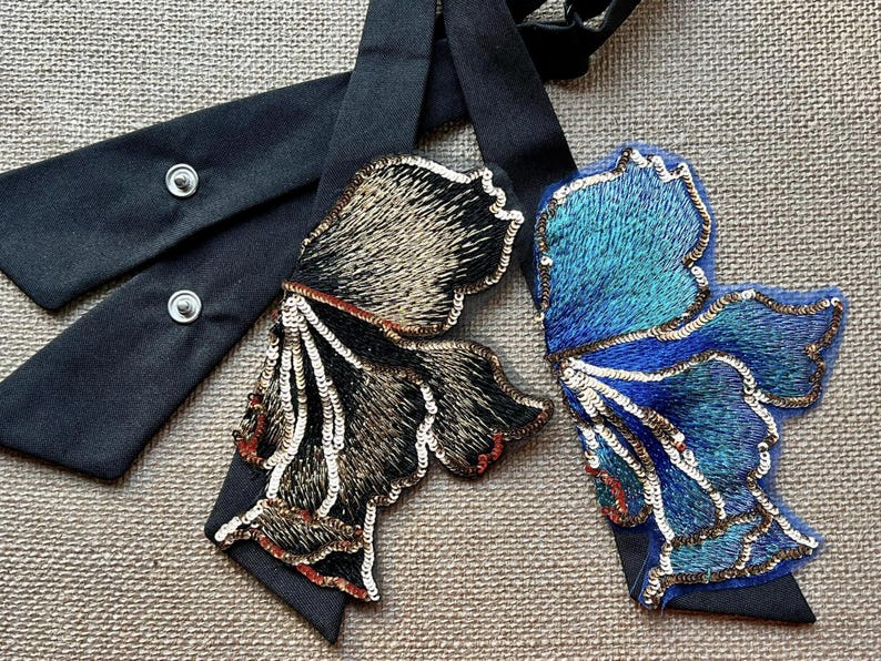 Puede incluir: Dos adornos decorativos con forma de mariposa con lazos de cinta negra. Una mariposa est&aacute; bordada con lentejuelas doradas, marrones y rojas. La otra es azul y turquesa con lentejuelas plateadas. Las cintas tienen cierres a presi&oacute;n.