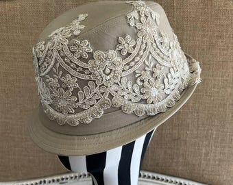 Könnte beinhalten: Taupefarbener Fedora-Hut mit weißem Spitzen-Overlay. Die Spitze zeigt Blumen- und Blättermuster, verziert mit kleinen Perlen. Der Hut hat eine strukturierte Krempe und wird auf einer schwarz-weiß gestreiften Form präsentiert.