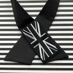 K&ouml;nnte beinhalten: Schwarze Krawatte mit Union Jack-Aufn&auml;her. Die Krawatte besteht aus einem glatten, dunklen Stoff und hat einen rechteckigen Aufn&auml;her mit einem wei&szlig;-schwarz gestickten britischen Flaggen-Design. Der Hintergrund ist schwarz-wei&szlig; gestreift.