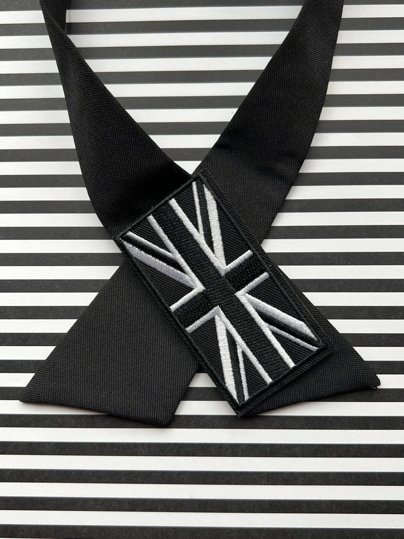 K&ouml;nnte beinhalten: Schwarze Krawatte mit Union Jack-Aufn&auml;her. Die Krawatte besteht aus einem glatten, dunklen Stoff und hat einen rechteckigen Aufn&auml;her mit einem wei&szlig;-schwarz gestickten britischen Flaggen-Design. Der Hintergrund ist schwarz-wei&szlig; gestreift.