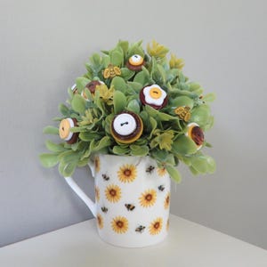 May include: A white ceramic mug decorated with yellow sunflower and bee patterns, filled with green artificial foliage. The foliage is accented with various round buttons in brown, white, and yellow, along with small wooden bee-shaped embellishments.