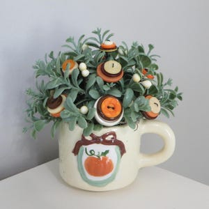 May include: A cream-colored ceramic mug featuring a raised pumpkin illustration and a brown bow design, filled with artificial green foliage and layered circular buttons in shades of orange, brown, and cream, accented with small white berries.