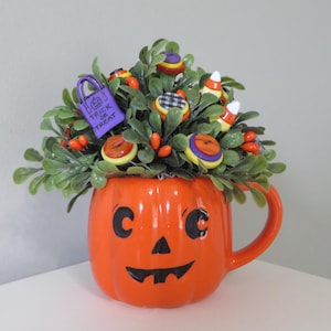 May include: An orange ceramic jack-o'-lantern mug filled with green faux foliage, colorful buttons, candy corn, and a purple charm shaped like a trick-or-treat bag with the text TRICK OR TREAT printed on the front in black lettering.
