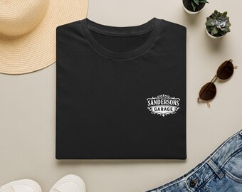 May include: A black t-shirt featuring a white graphic on the chest with the text SANDERSONS GARAGE enclosed in a decorative border. The shirt is displayed alongside a straw hat, two small potted succulents, brown sunglasses, and blue jeans.