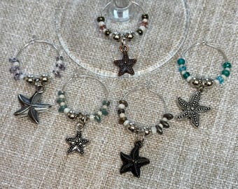 May include: Six silver wine glass charms with beaded accents and starfish pendants. The charms are in various colors, including white, blue, green, and brown.